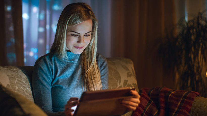 Beautiful Young Woman in Her Living Room. She is Sittin on a Sofa and Uses Tablet Computer. Behind Her Big City is Seen in the Window. Cold ambient.